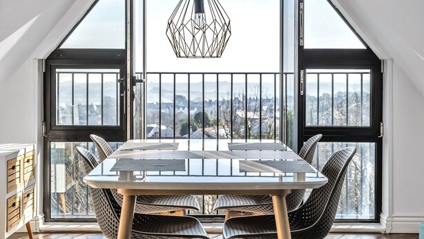A dining area with a table and chairs, a hanging light fixture, and large windows with a view of the city. - Pochin House Collections (St Austell)