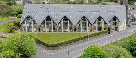 A large, modern building with a symmetrical roofline and multiple windows, surrounded by greenery and a road.