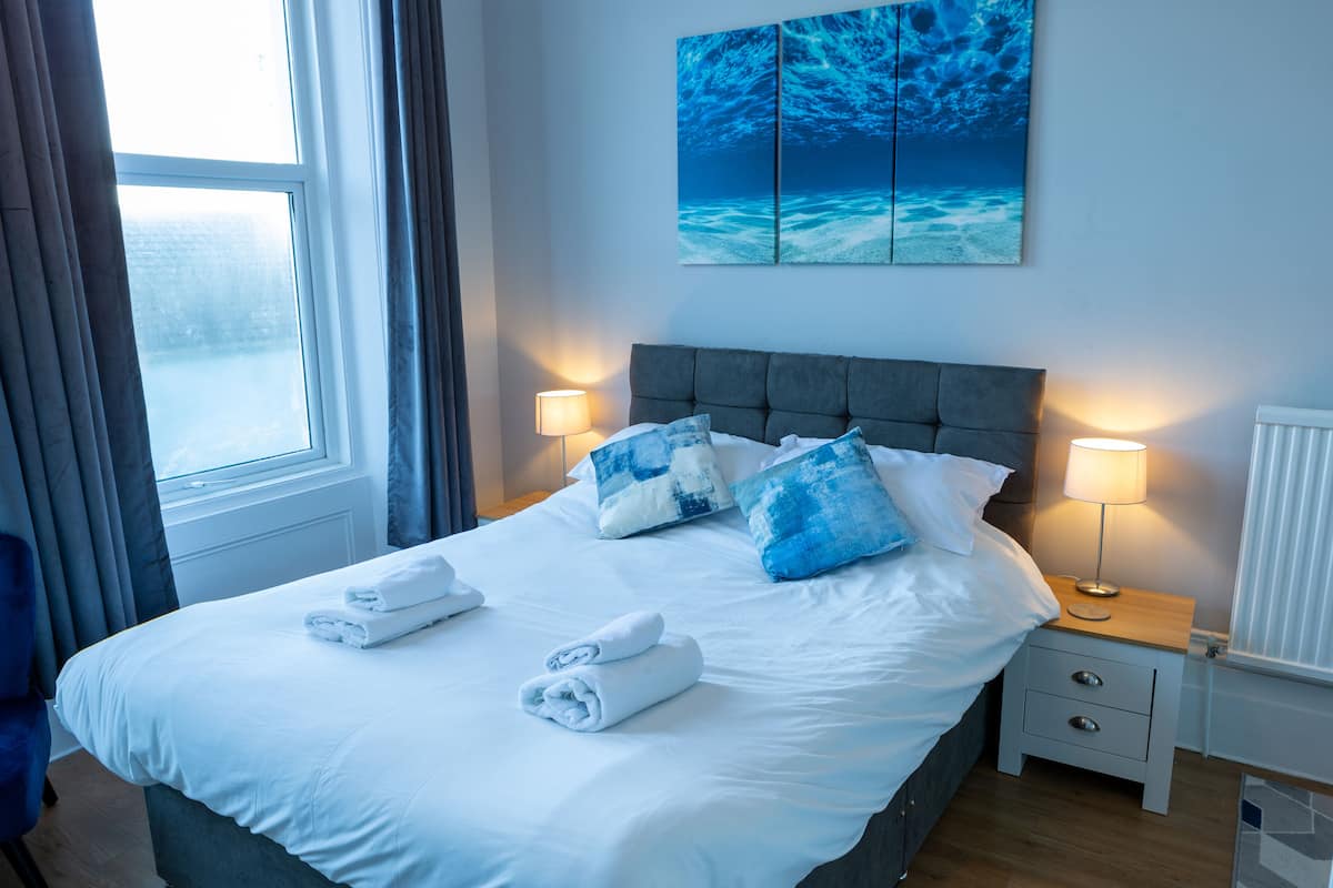 A neatly made bed with white linens and blue pillows, a bedside table with a lamp, and a large ocean-themed painting on the wall.