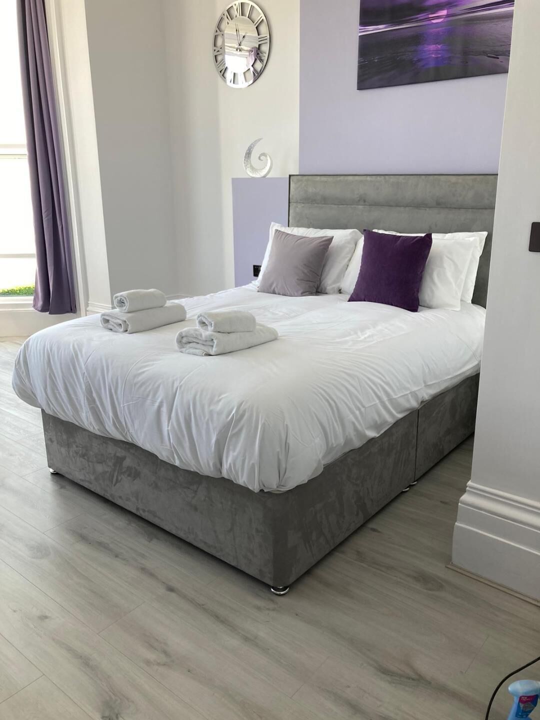 Une chambre à coucher moderne avec une tête de lit grise, du linge de lit blanc et des serviettes soigneusement pliées sur le lit.