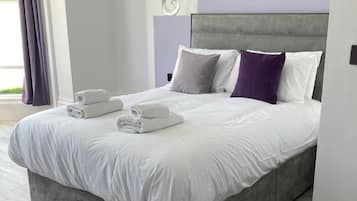 Une chambre à coucher moderne avec une tête de lit grise, du linge de lit blanc et des serviettes soigneusement pliées sur le lit.