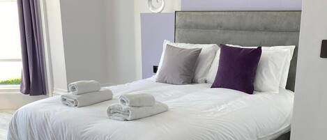 A modern bedroom with a grey headboard, white bedding, and neatly folded towels on the bed.