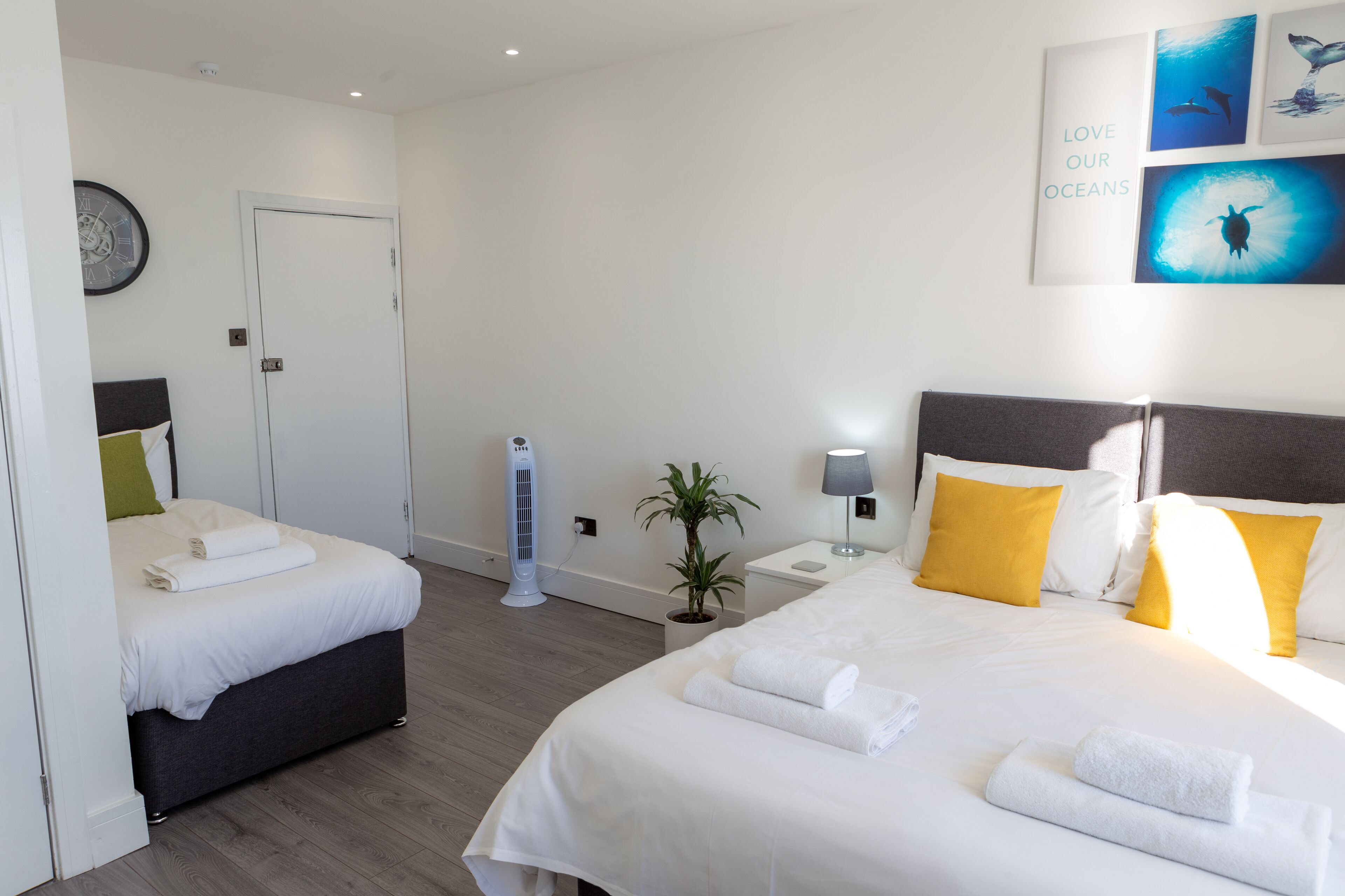 A hotel room with two beds, a bedside table with a lamp, a wall clock, and framed pictures.