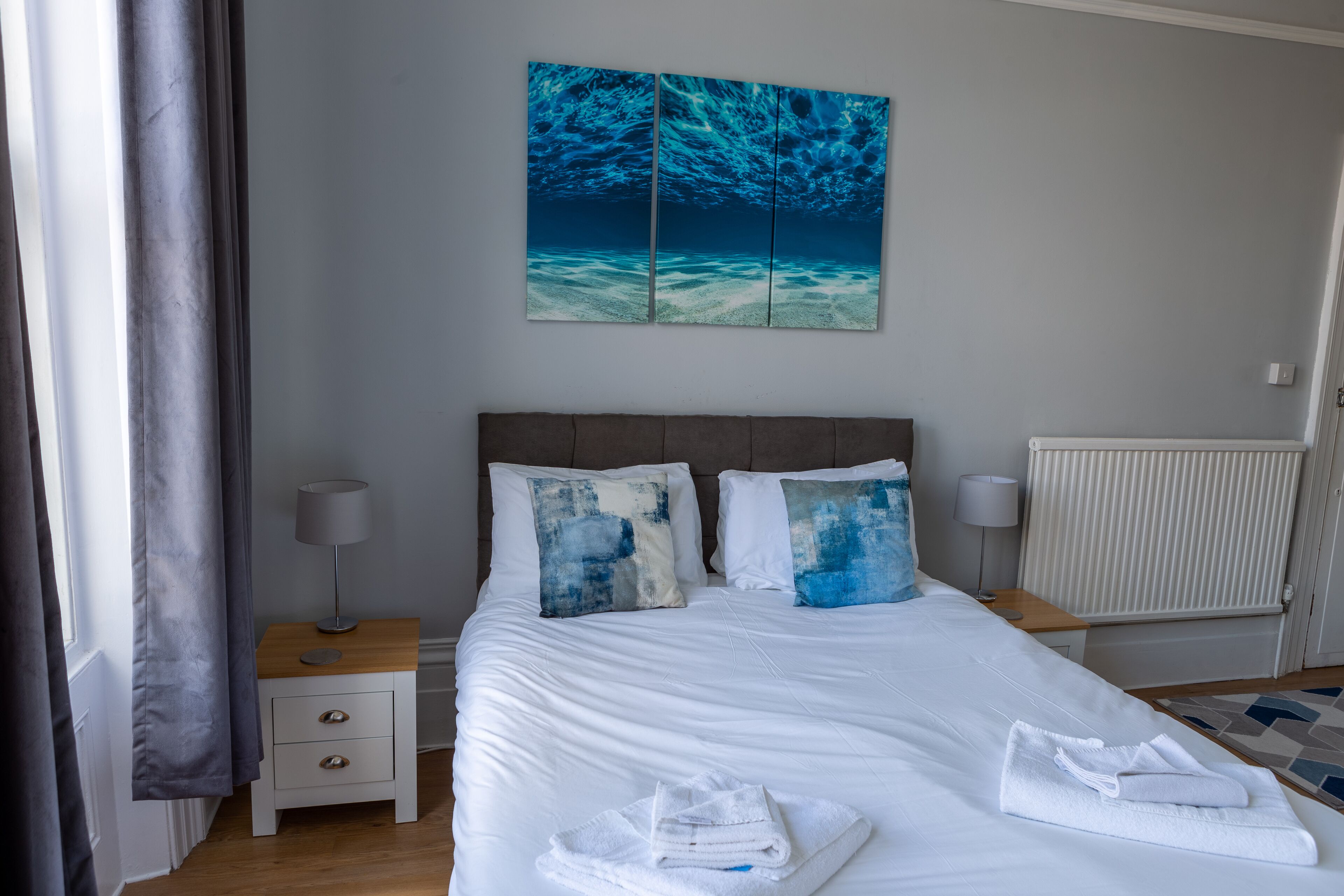 A neatly made bed with white linens, two pillows with abstract designs, a bedside table with a lamp, and a painting of an underwater scene above the bed.