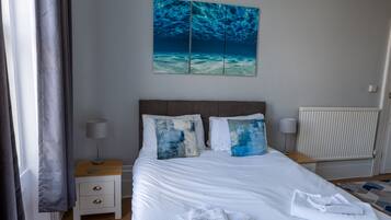 A neatly made bed with white linens, two pillows with abstract designs, a bedside table with a lamp, and a painting of an underwater scene above the bed.