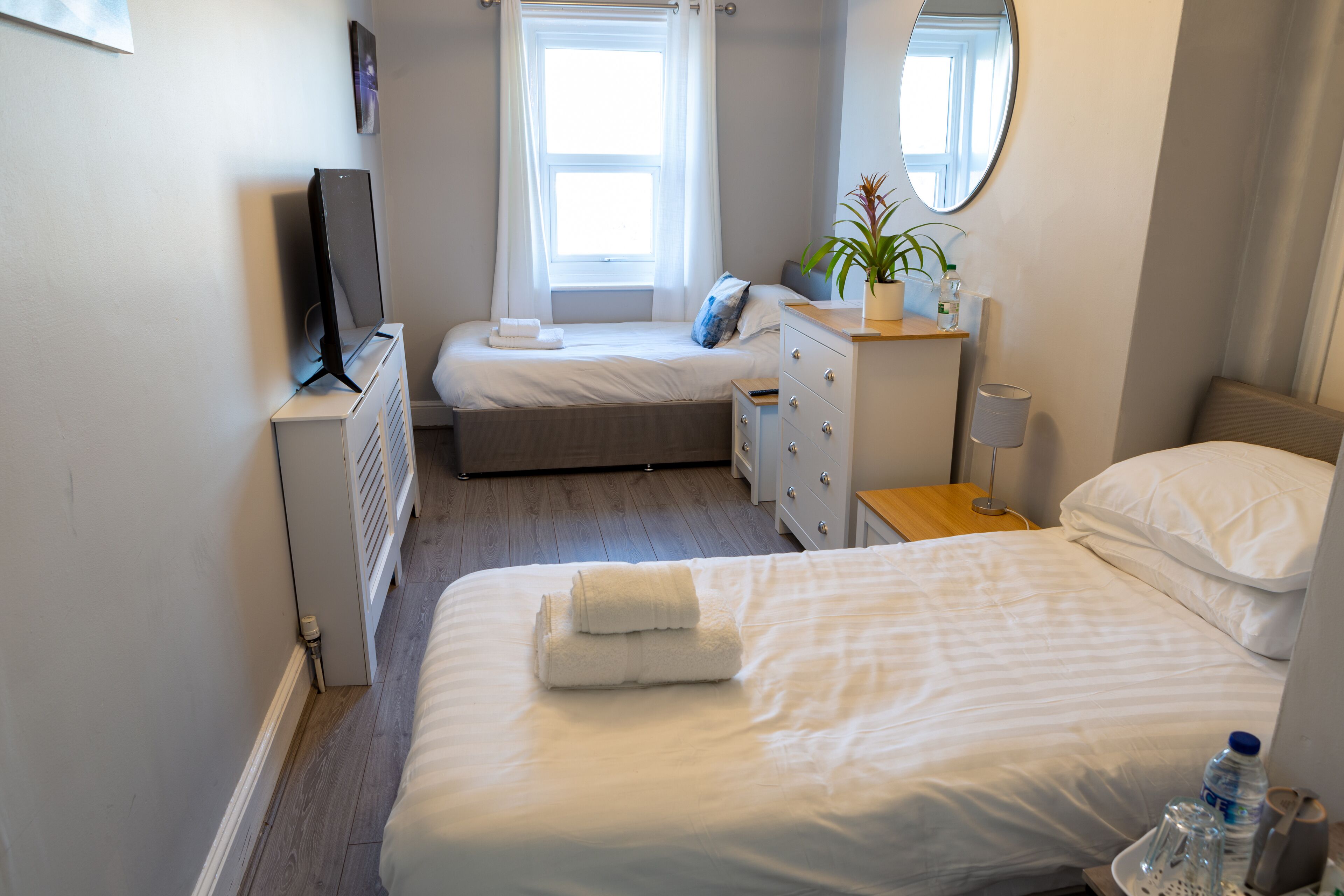 Hotellihuone, jossa on sänky, yöpöytä lampun kanssa, lipasto, televisio ja ikkuna, jossa on verhot.