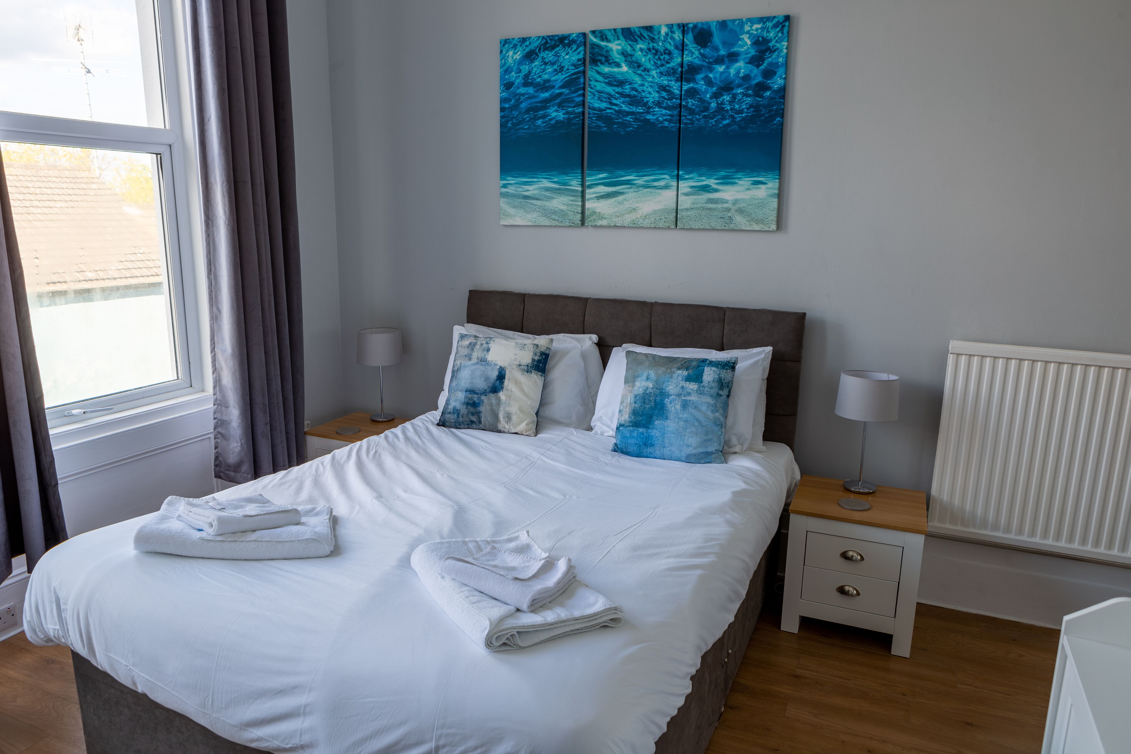 A neatly made bed with white linens and blue pillows, a bedside table with a lamp, and a large window with curtains.