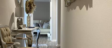 Uma sala de estar ampla, com uma poltrona grande, uma mesinha de apoio com abajur, e um espelho decorativo.
