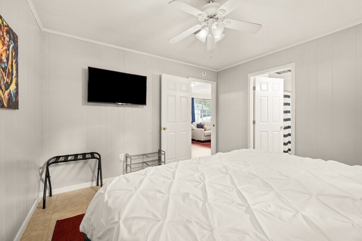 A bedroom with a large bed, a ceiling fan, a TV mounted on the wall, and a door leading to another room.