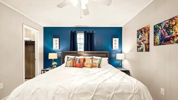 A bedroom with a large bed, two bedside tables with lamps, a ceiling fan, a window with curtains, and two wall-mounted paintings.