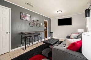 Una sala de estar moderna con un sofá gris, un otoman negro y un tapete rojo y negro. Hay una bicicleta montada en la pared y un televisor de pantalla plana sobre el sofá.