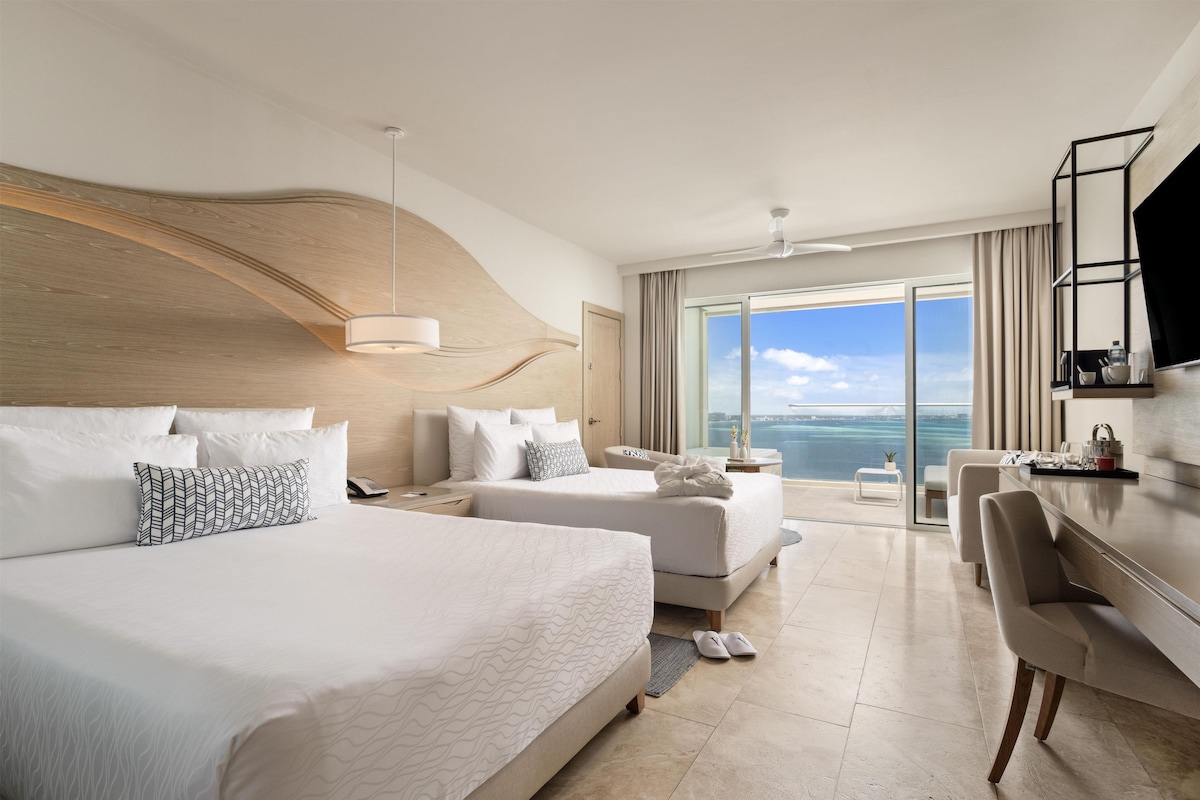 A hotel room with two beds, a desk, and a large window offering a view of the sea.