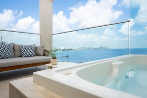 A balcony with a sofa and a hot tub overlooking a body of water with buildings in the distance.