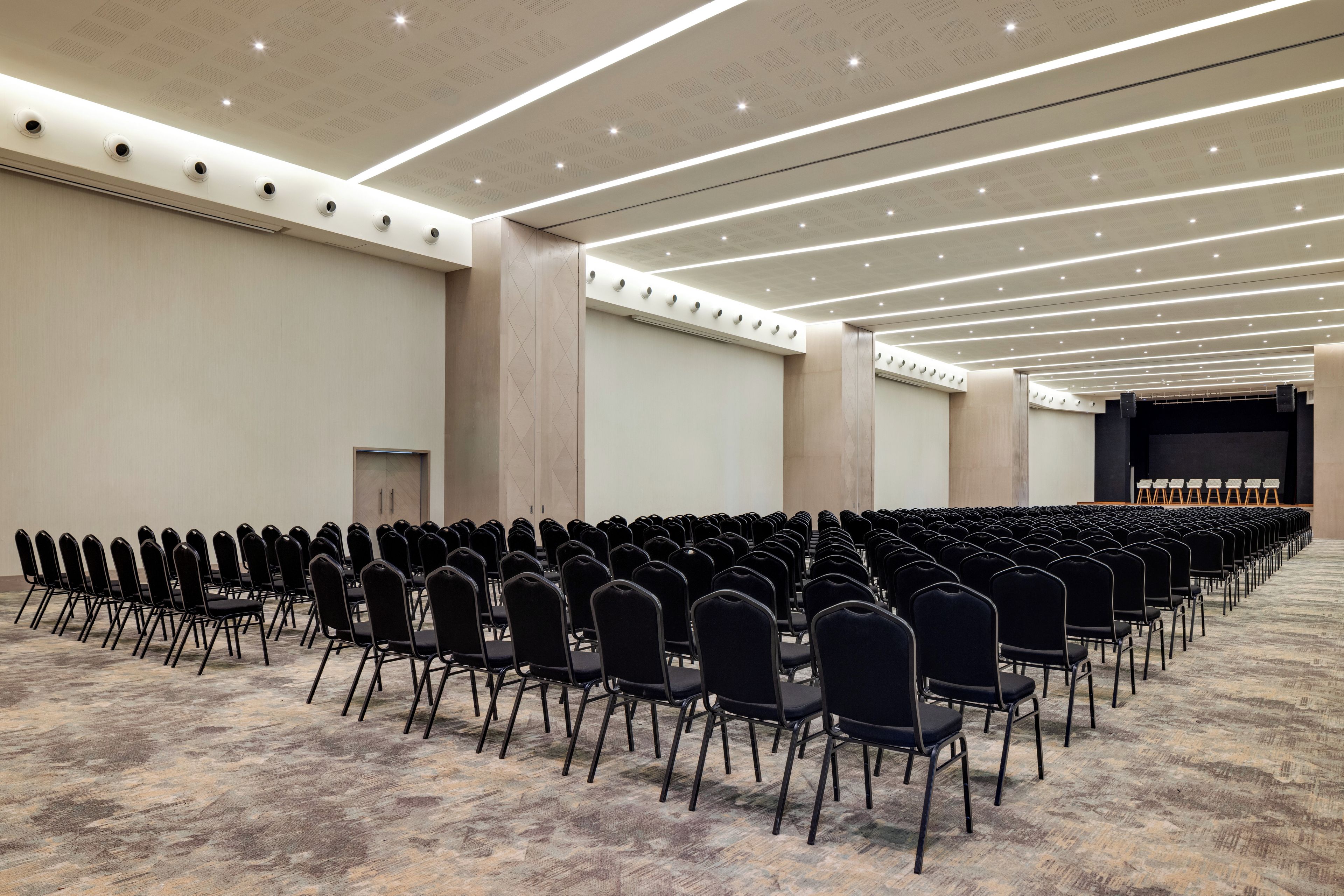 Un salón de conferencias con filas de sillas, un escenario e iluminación en el techo.