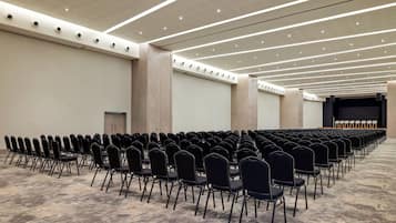 Un salón de conferencias con filas de sillas, un escenario e iluminación en el techo.