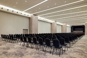 Un salón de conferencias con filas de sillas, un escenario e iluminación en el techo.