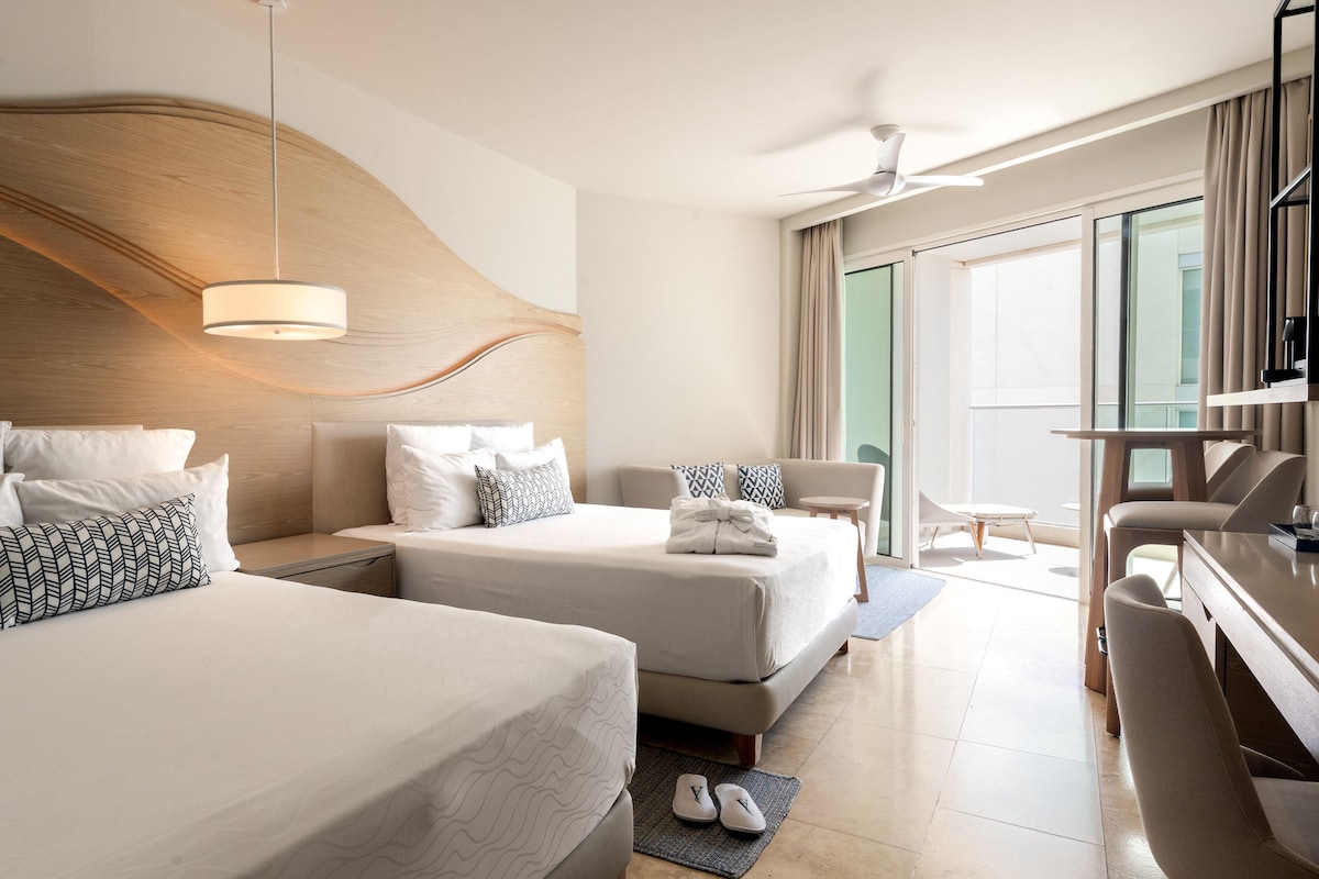 A hotel room with two beds, a desk, a chair, a TV, and a balcony with a view.