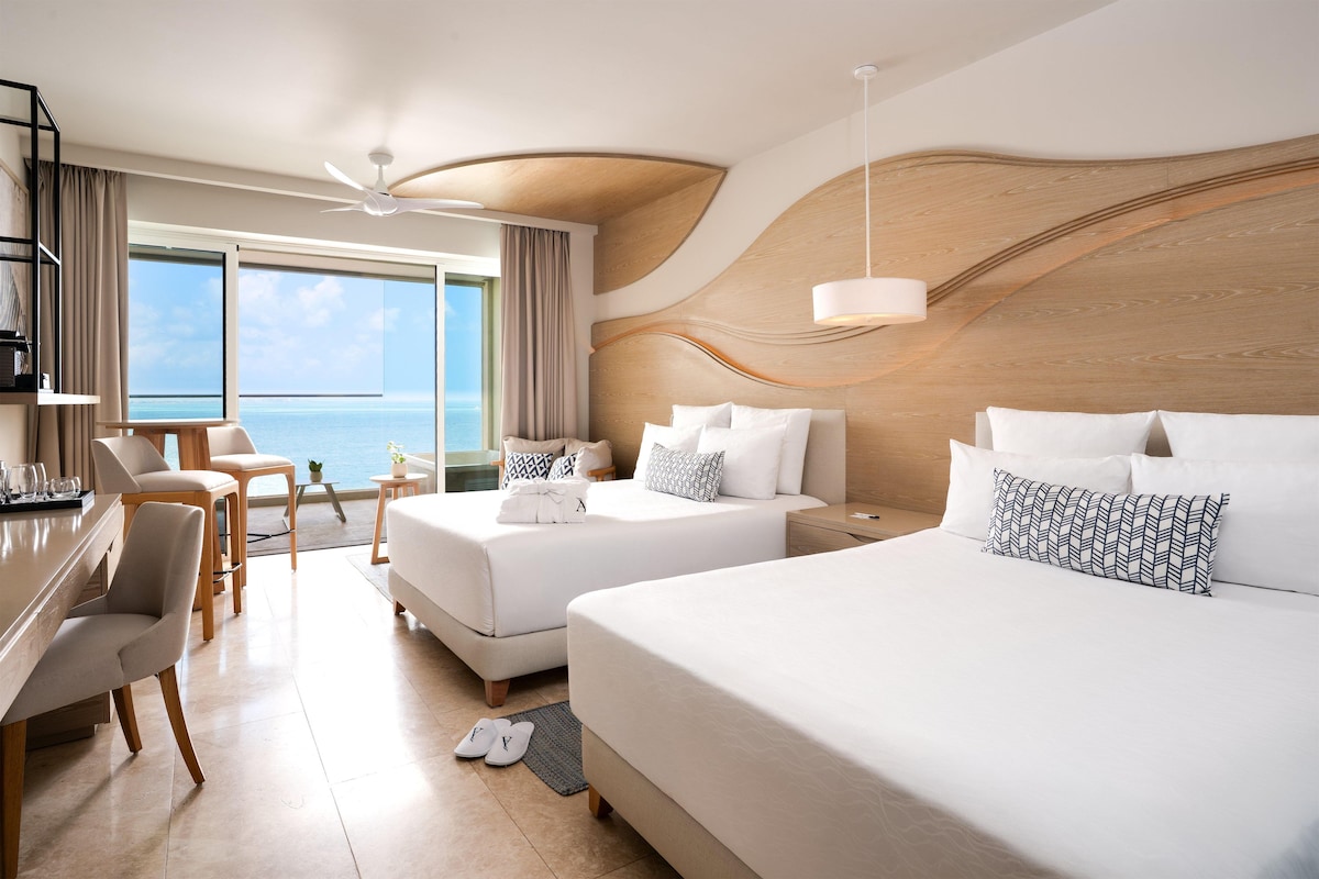A hotel room with two beds, a desk, a chair, a view of the sea, and a ceiling fan.