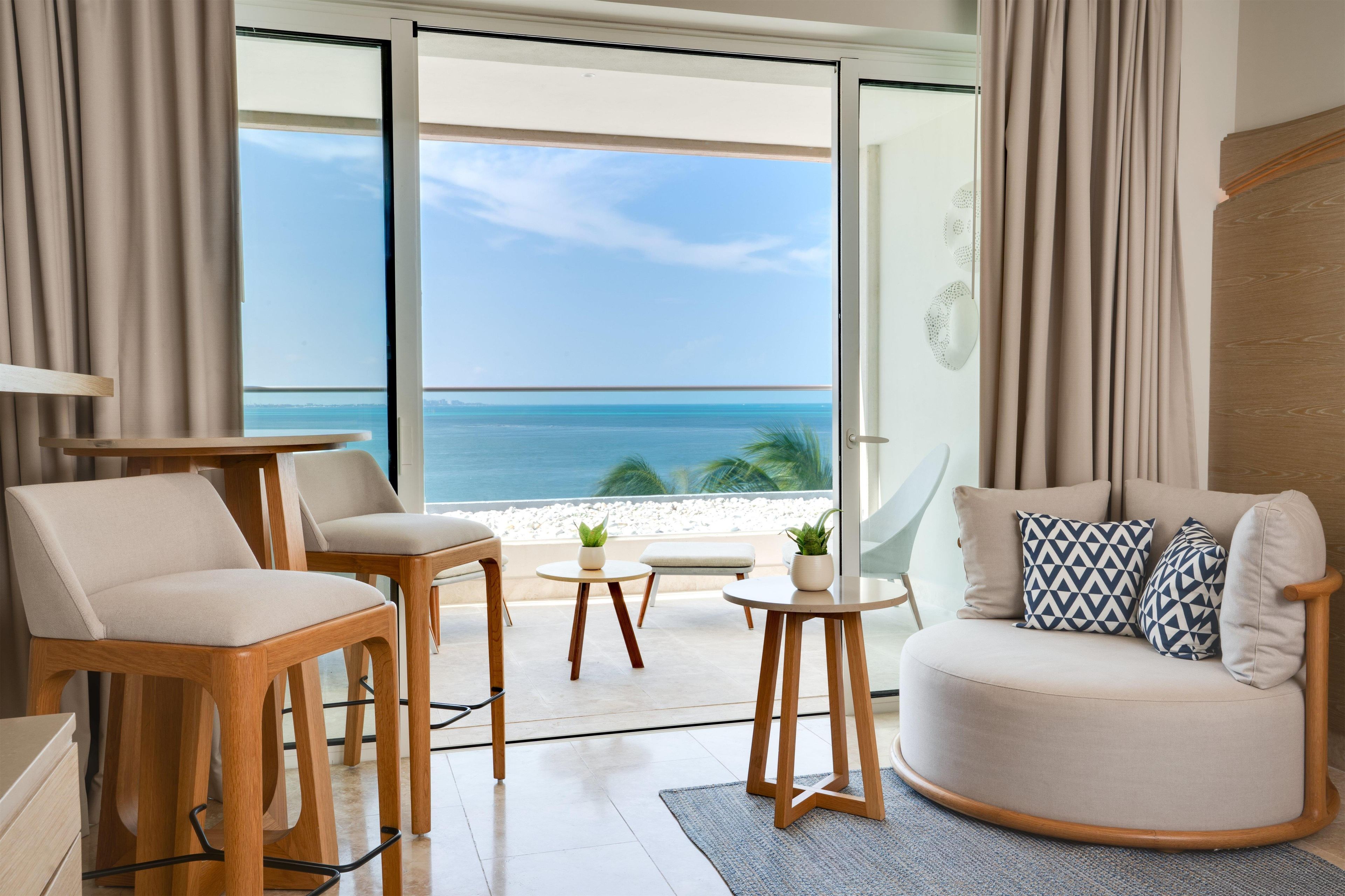 Uma sala de estar moderna com vista para o oceano, com um sofá, cadeiras, uma mesa pequena e um vaso de planta.