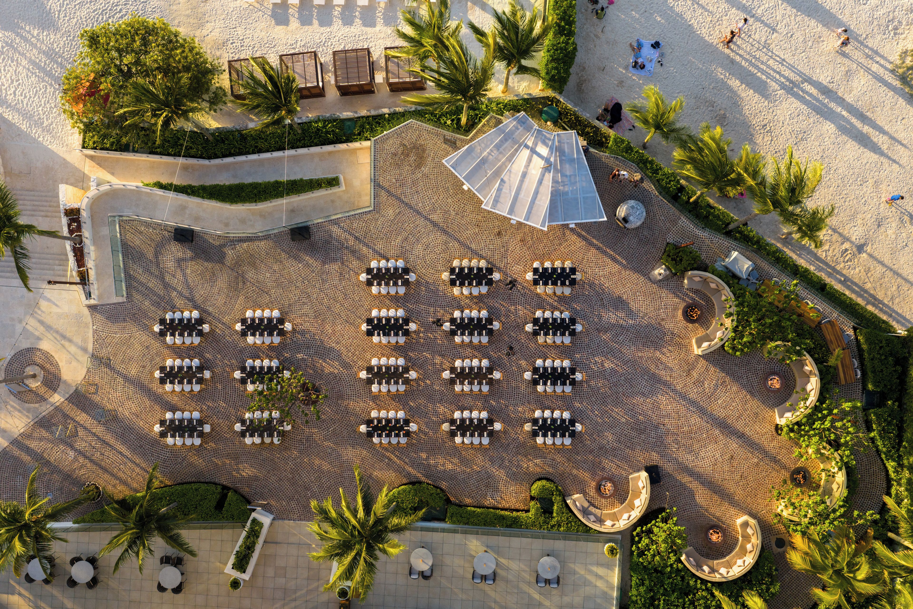 Vista aérea de un hotel con varias zonas de estar al aire libre, incluyendo mesas redondas y tumbonas, rodeado de palmeras y una playa de arena.