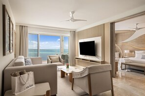 A modern hotel room with a large TV, comfortable seating, a view of the sea, and a ceiling fan.