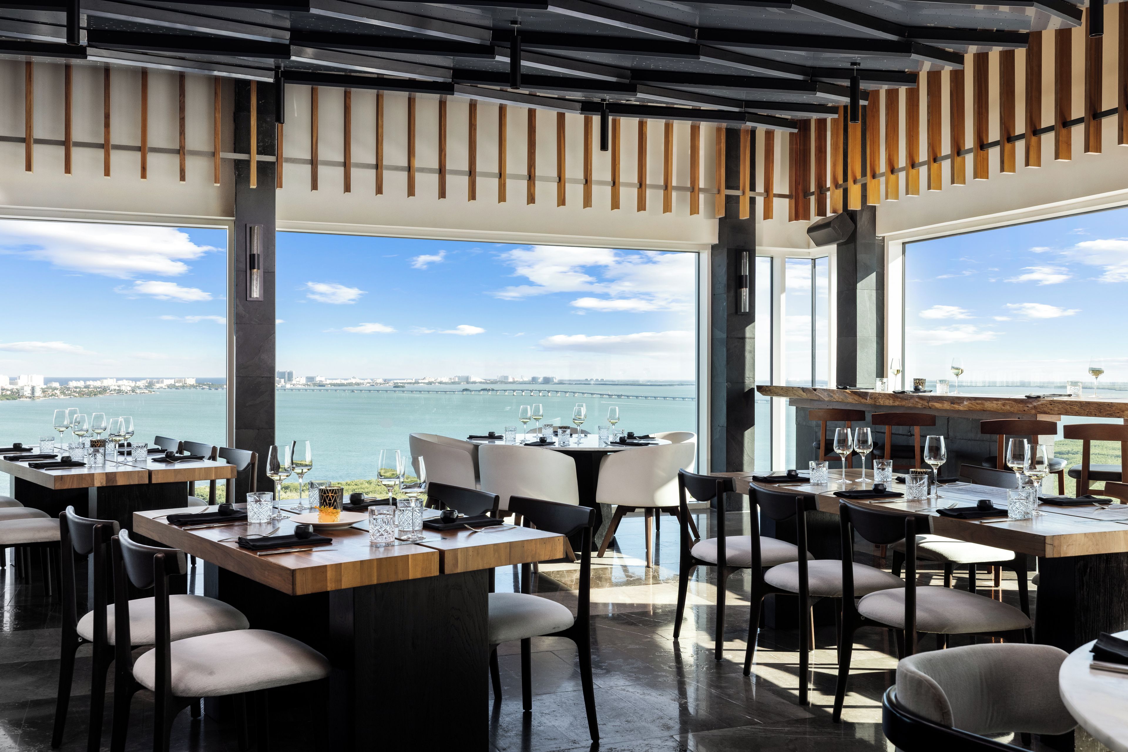 A restaurant with multiple tables set for dining, large windows offering a view of the water and city skyline, and a modern interior design with wooden beams and metal accents.