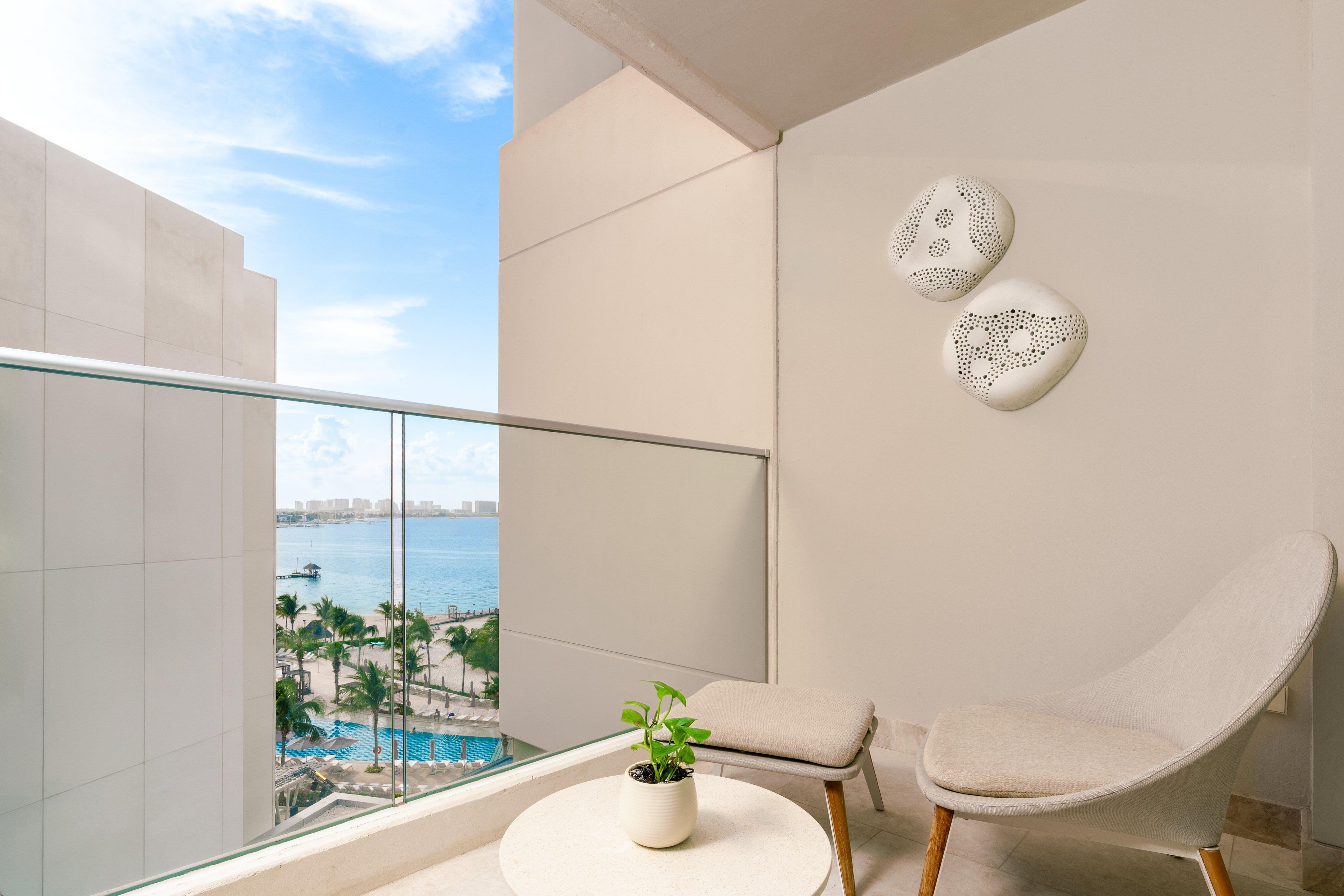 A balcony with a glass railing, a potted plant, a beige chair, and two wall art pieces.