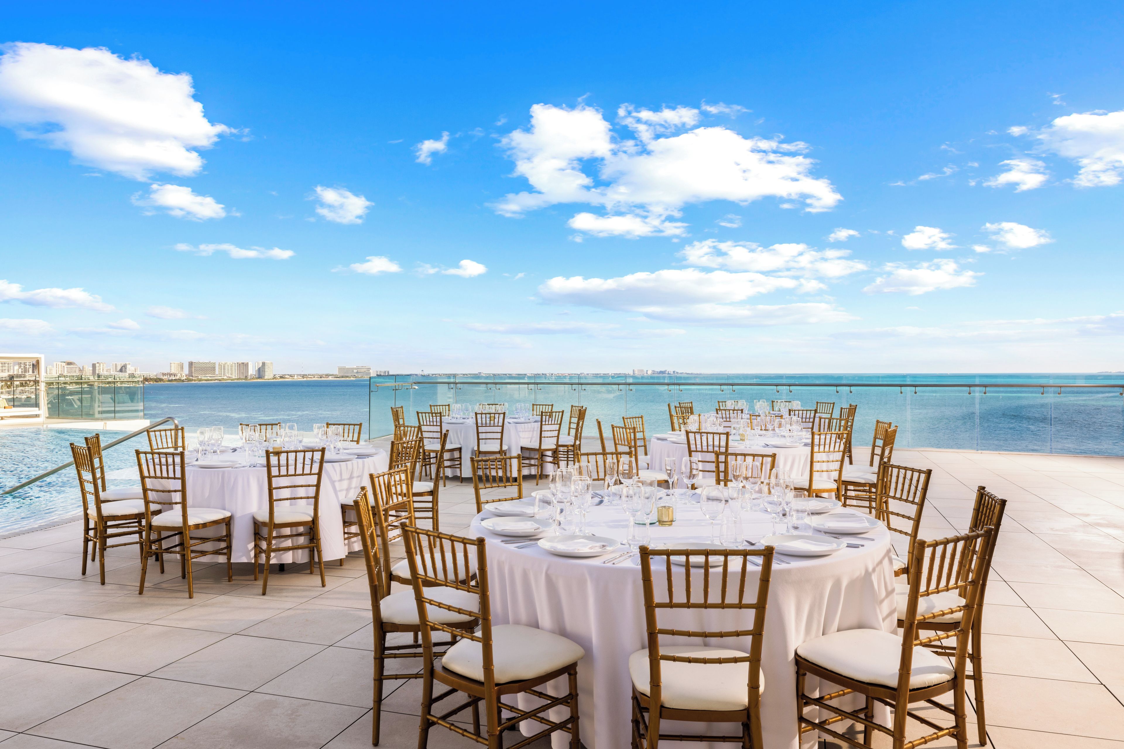 Un área de comedor en la azotea con mesas redondas dispuestas para una comida, con vista a una masa de agua y el horizonte de una ciudad.