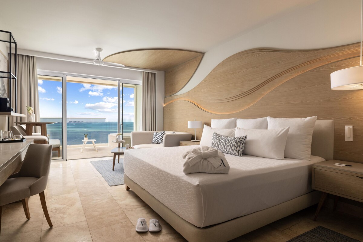 A modern hotel room with a large bed, a desk, and a view of the sea.