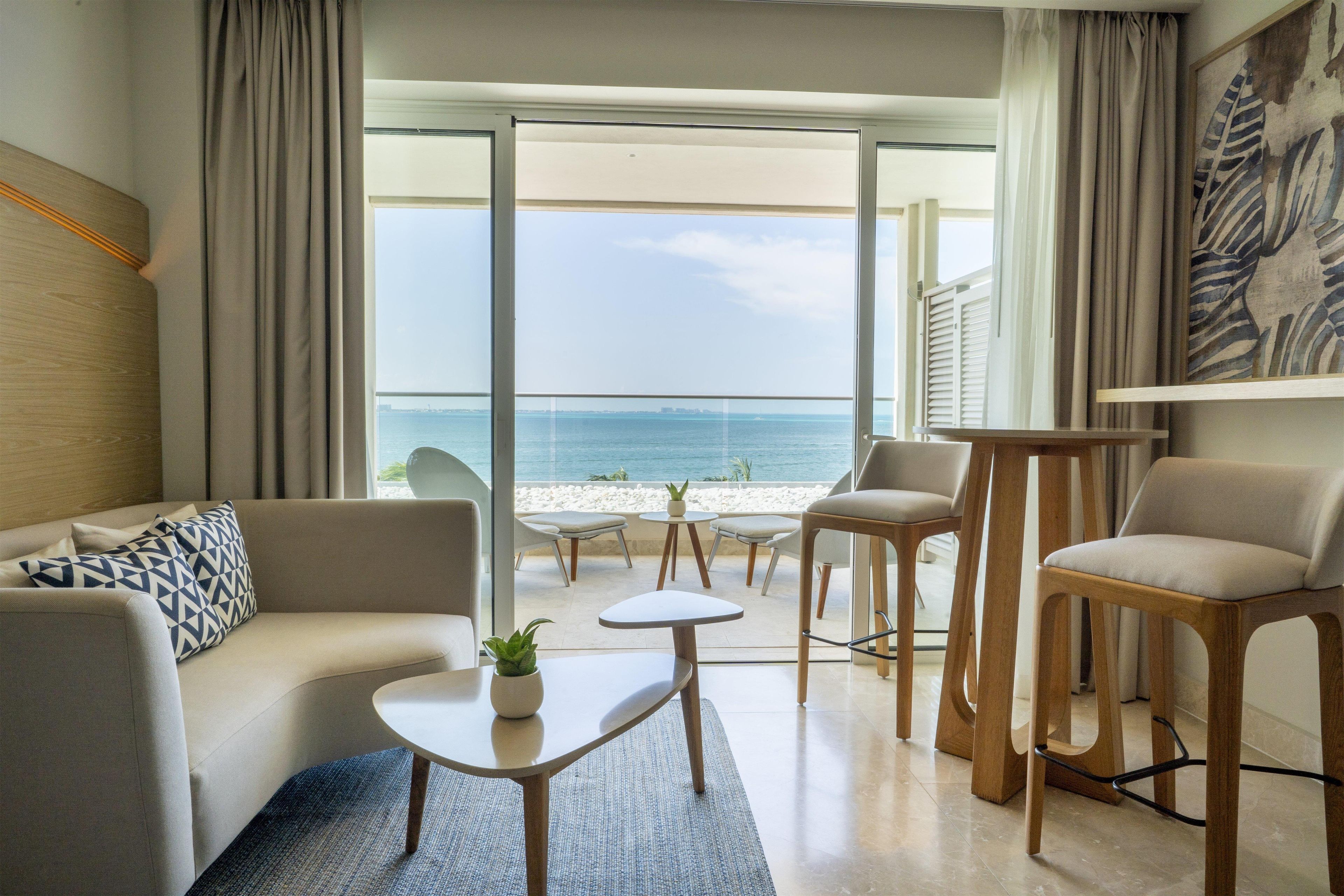 A modern hotel room with a sofa, two chairs, a small table, and a view of the sea.