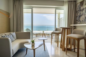 Quarto de hotel moderno com sofá, duas cadeiras, uma mesinha e vista para o mar.