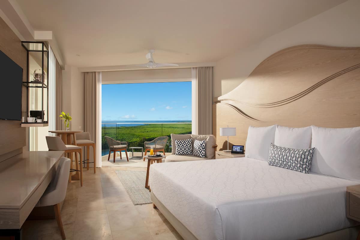 Een hotelkamer met een groot bed, een bureau, een stoel, een tafeltje en uitzicht op een groen landschap door een groot raam.