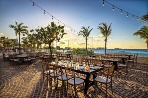 An outdoor dining area with tables and chairs, palm trees, and a view of the sea.