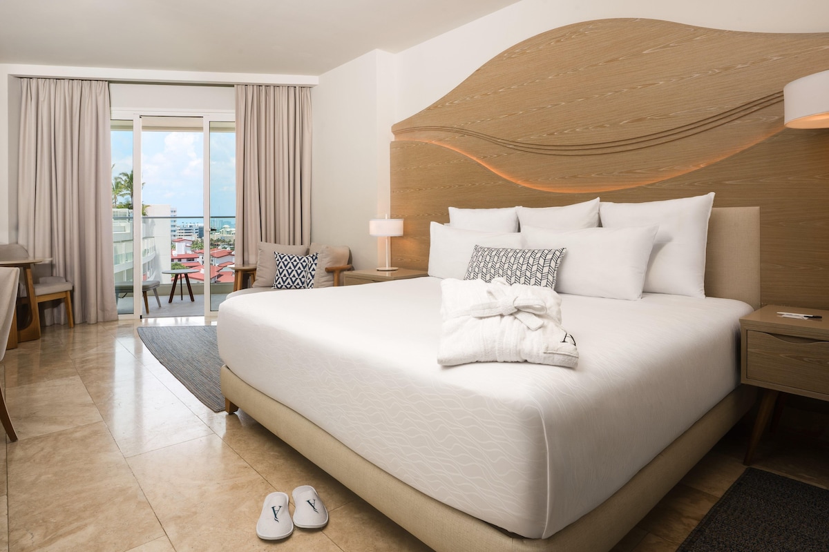 A hotel room with a large bed, a nightstand, a chair, a desk, and a view of the city through the window.