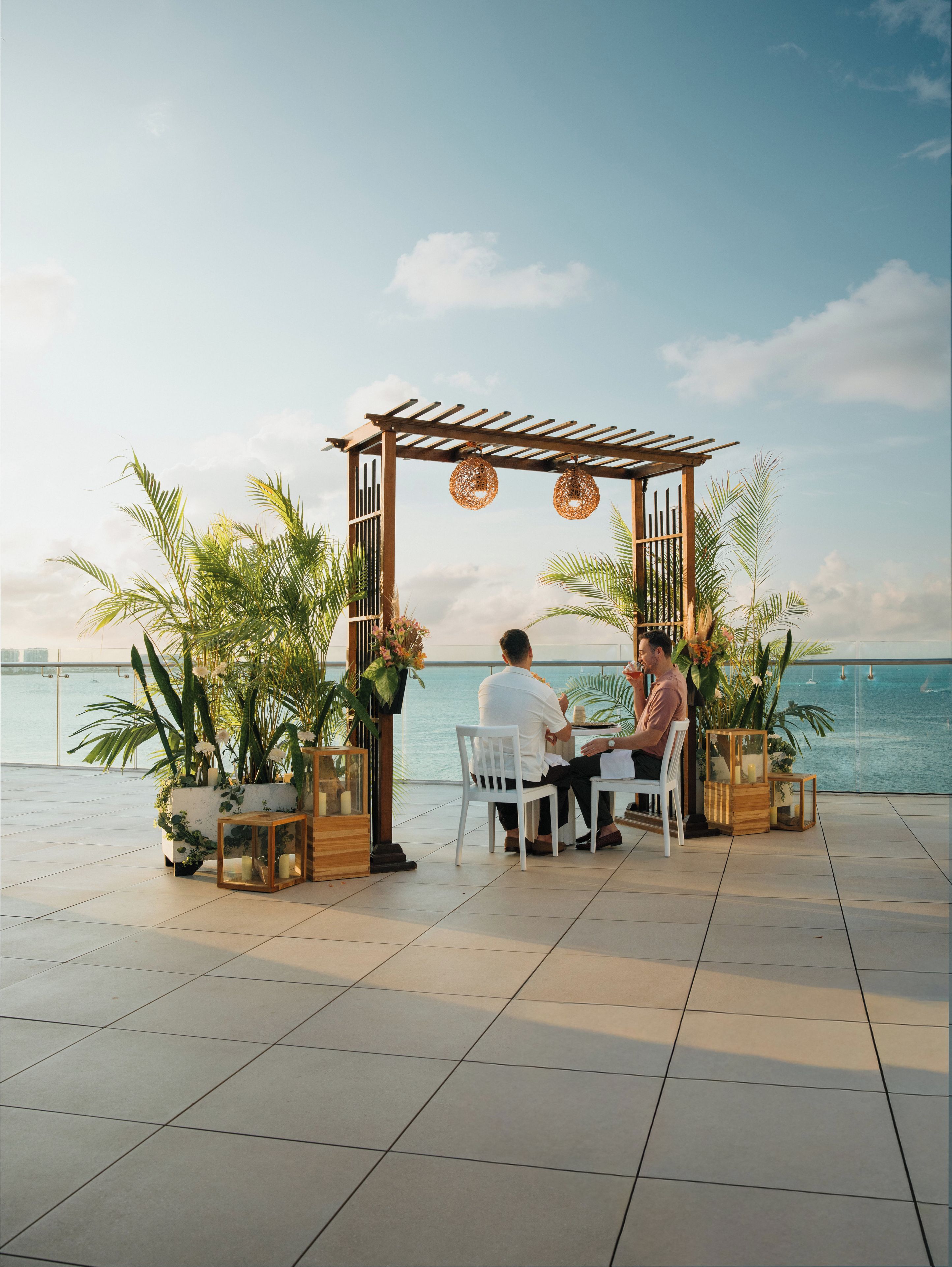 A couple seated at a table under a wooden pergola with a view of the sea.