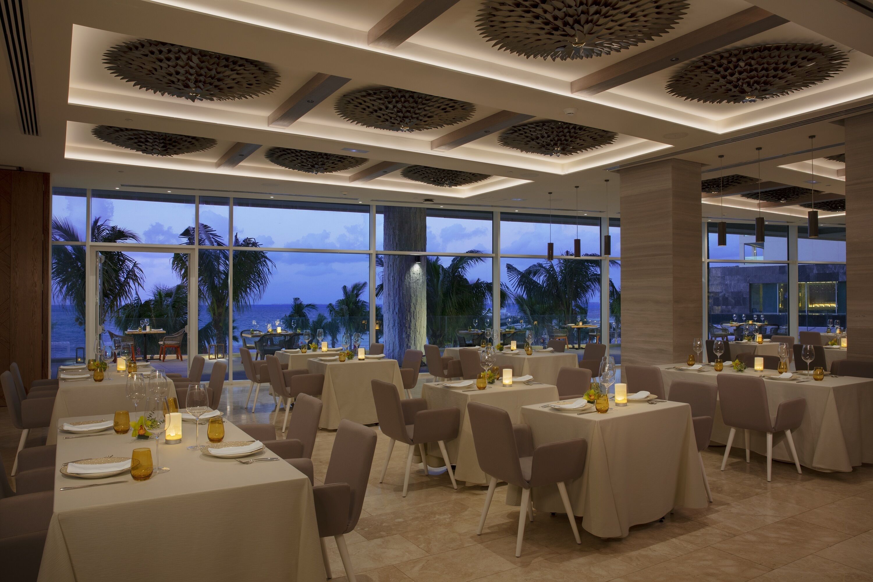 Restaurante com mesas preparadas para refeições, grandes janelas com vista para o mar e design moderno no teto.