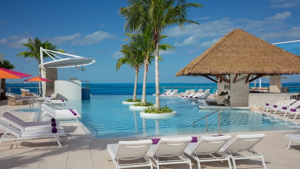 Un espace aménagé au bord de la piscine, avec des chaises longues, des palmiers et une structure dotée d’un toit de chaume.