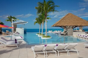 Un espace aménagé au bord de la piscine, avec des chaises longues, des palmiers et une structure dotée d’un toit de chaume.