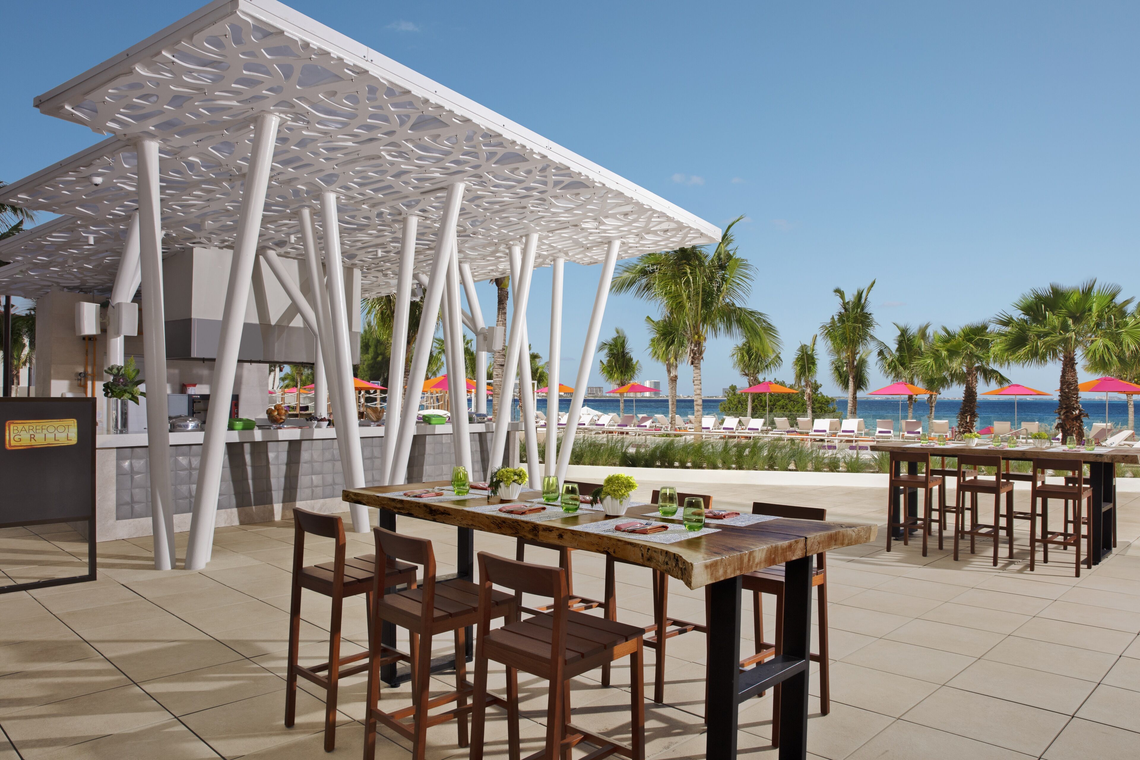 Zona pranzo all'aperto con una pergola bianca, tavoli e sedie di legno, affacciata su una piscina con palme e ombrelloni.
