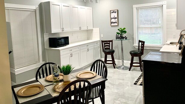 A modern kitchen with white cabinets, a black microwave, and a dining area with a glass table and chairs. - The Roulette Duplex-Private Yard&Parking (Niagara Falls)