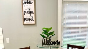 A dining area with two wooden chairs, a glass table, and a 'Welcome' sign with a plant. - The Roulette Duplex-Private Yard&Parking (Niagara Falls)