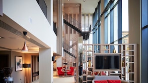 Een ruime hotellobby met een hoge plafond, een trap, een zithoek met rode stoelen en een groot televisiescherm.