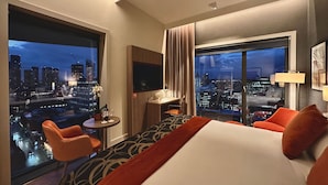 Hotelkamer met een groot bed, een bureau met stoel, een televisie en uitzicht op een stadsbeeld 's nachts.