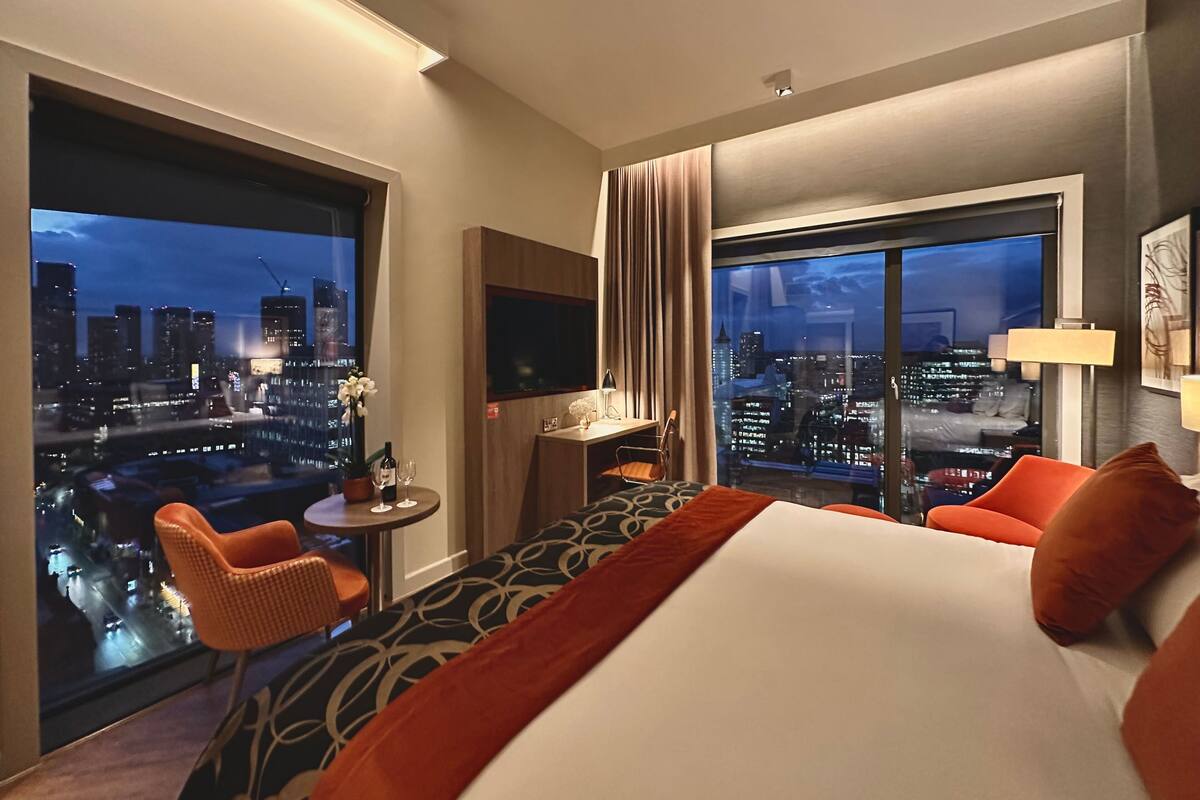 Une chambre d’hôtel avec un grand lit, un bureau avec une chaise, une télévision et une vue sur le paysage urbain illuminé la nuit.