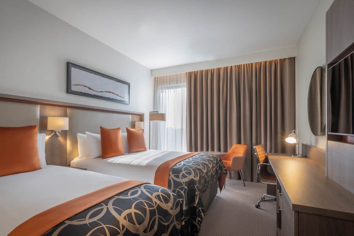 Une chambre d’hôtel avec deux lits, un bureau, une chaise et une grande fenêtre avec des rideaux.