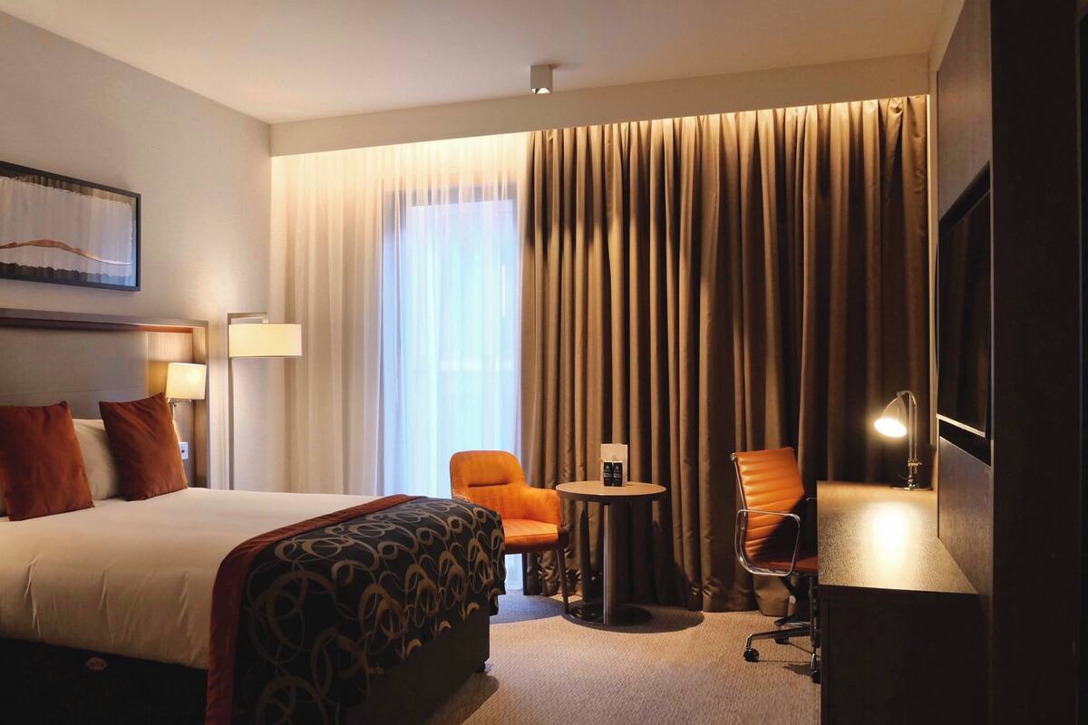 Une chambre d’hôtel comprenant un lit, deux tables de chevet avec des lampes, un bureau avec une chaise, une petite table ronde avec une chaise, et une grande fenêtre avec des rideaux.