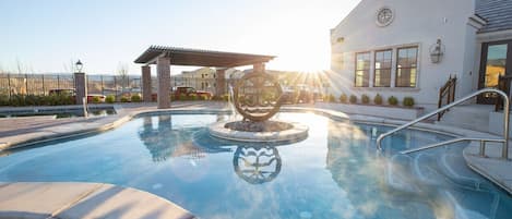 Una piscina con una escultura decorativa en el centro, una zona de estar cubierta y un edificio con amplias ventanas y una puerta.