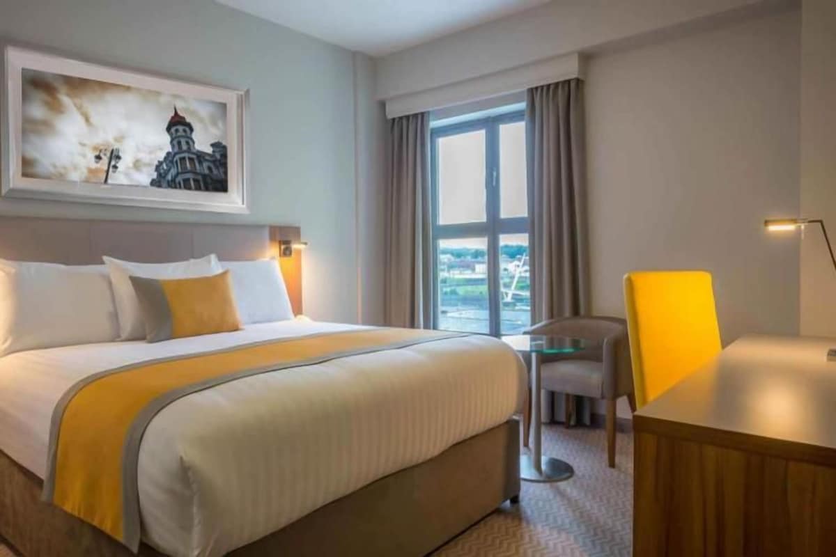 A hotel room with a bed, a desk, a chair, a window with a view, and a painting on the wall.