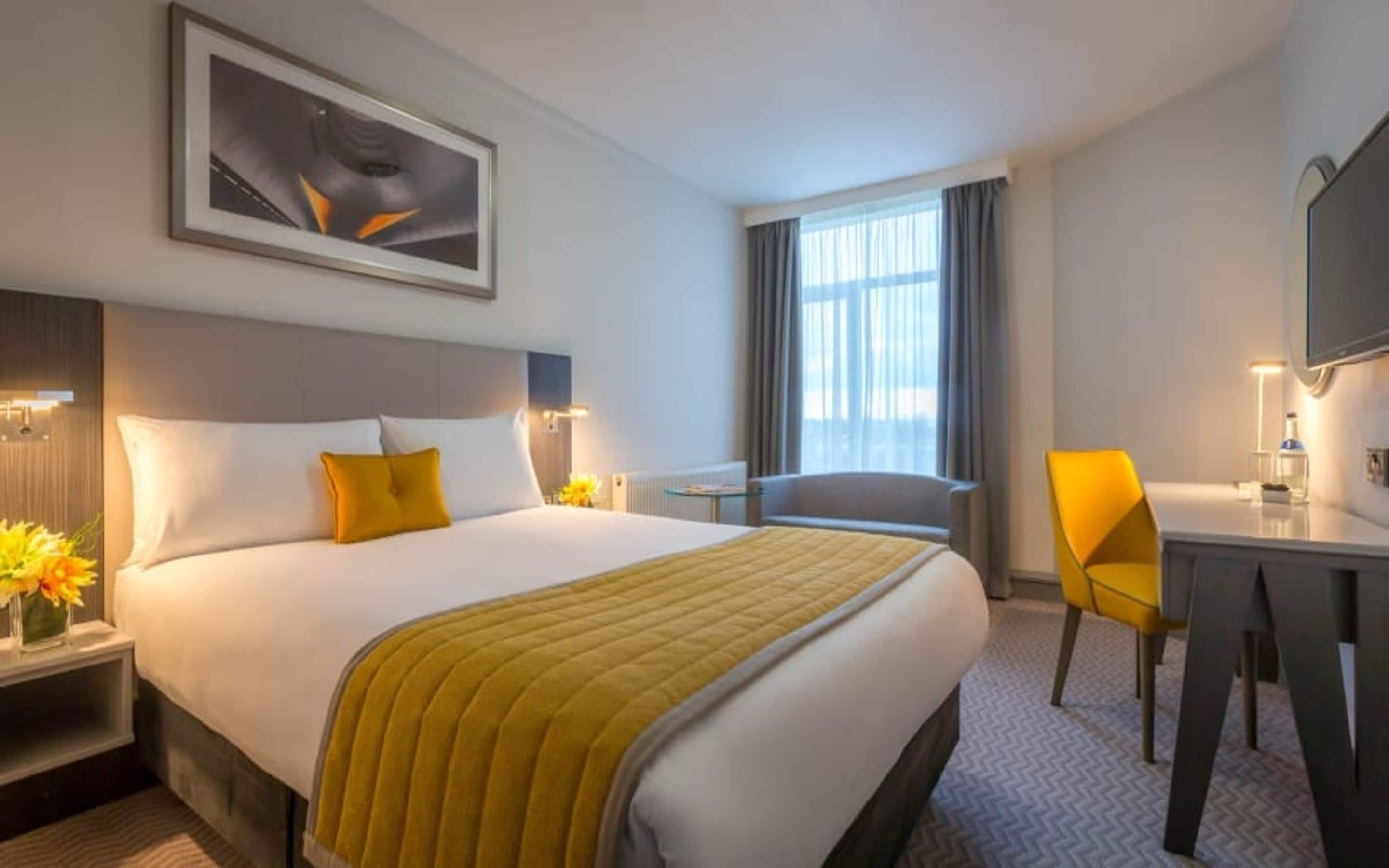 Habitación de hotel con cama, escritorio, silla, ventana y un cuadro en la pared.