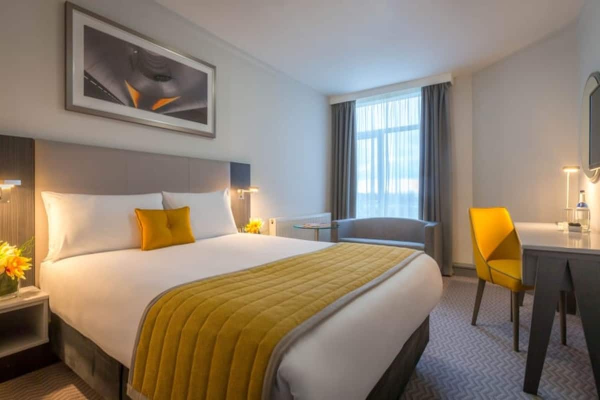 A hotel room with a bed, a desk, a chair, a window, and a painting on the wall.